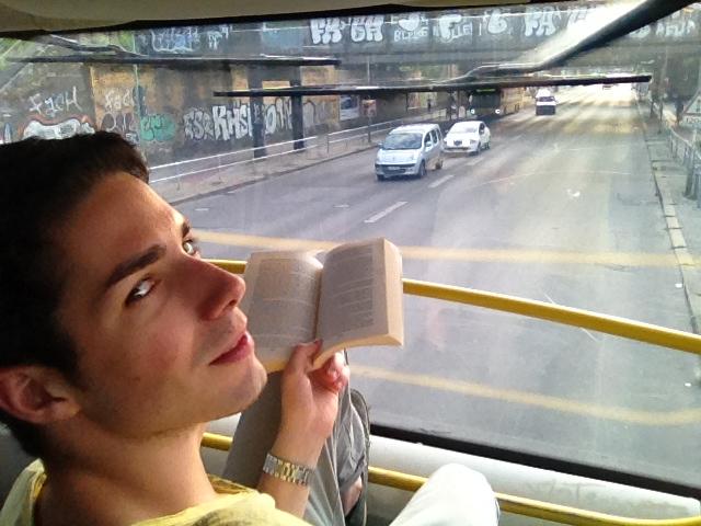 Tiziano Boccaccini al piano superiore di un autobus scoperto, con un libro aperto in mano, si volta verso l'obiettivo