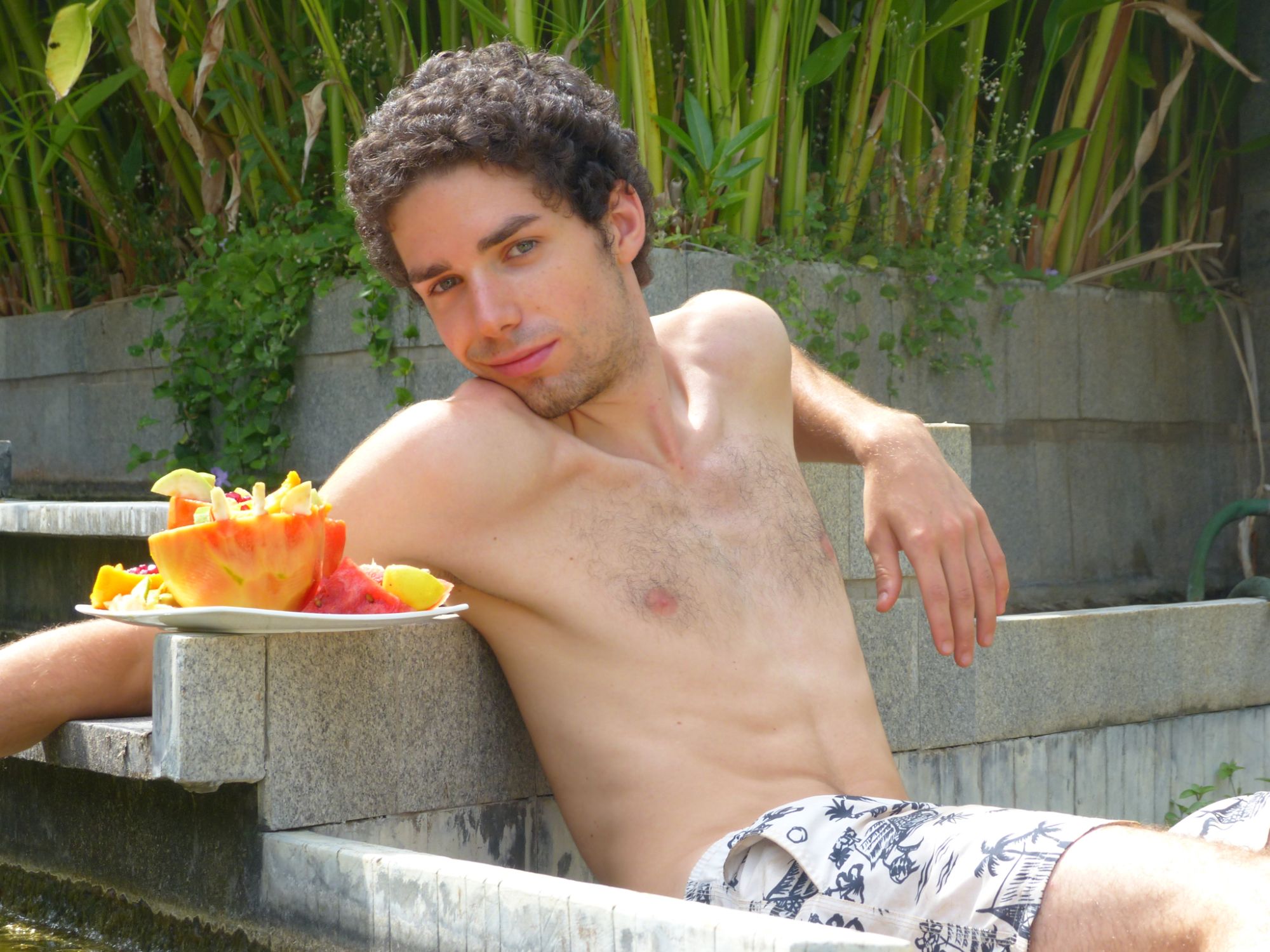 Tiziano Boccaccini a torso nudo appoggiato al bordo di una vasca in granito, accanto a un piatto di frutta tropicale, con piante verdi sullo sfondo