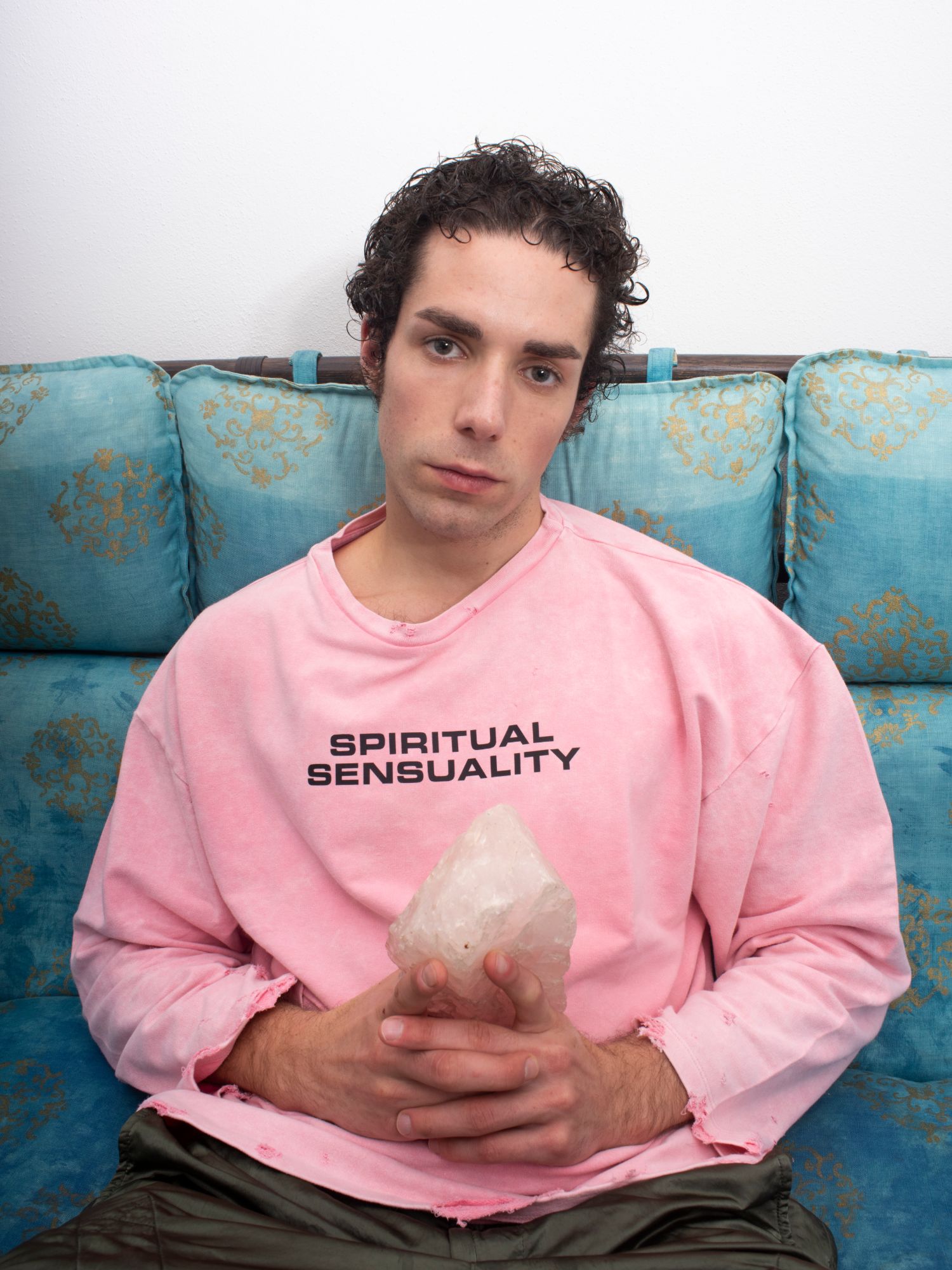 Tiziano Boccaccini seduto su un divano azzurro con cuscini decorati in oro, in felpa rosa con la scritta Spiritual Sensuality e un cristallo di quarzo rosa tra le mani